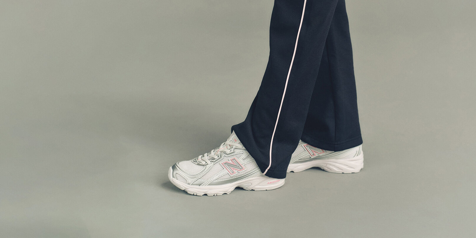 Close-up studio image of the lower legs of someone wearing New Balance sneakers against a plain light gray background.
