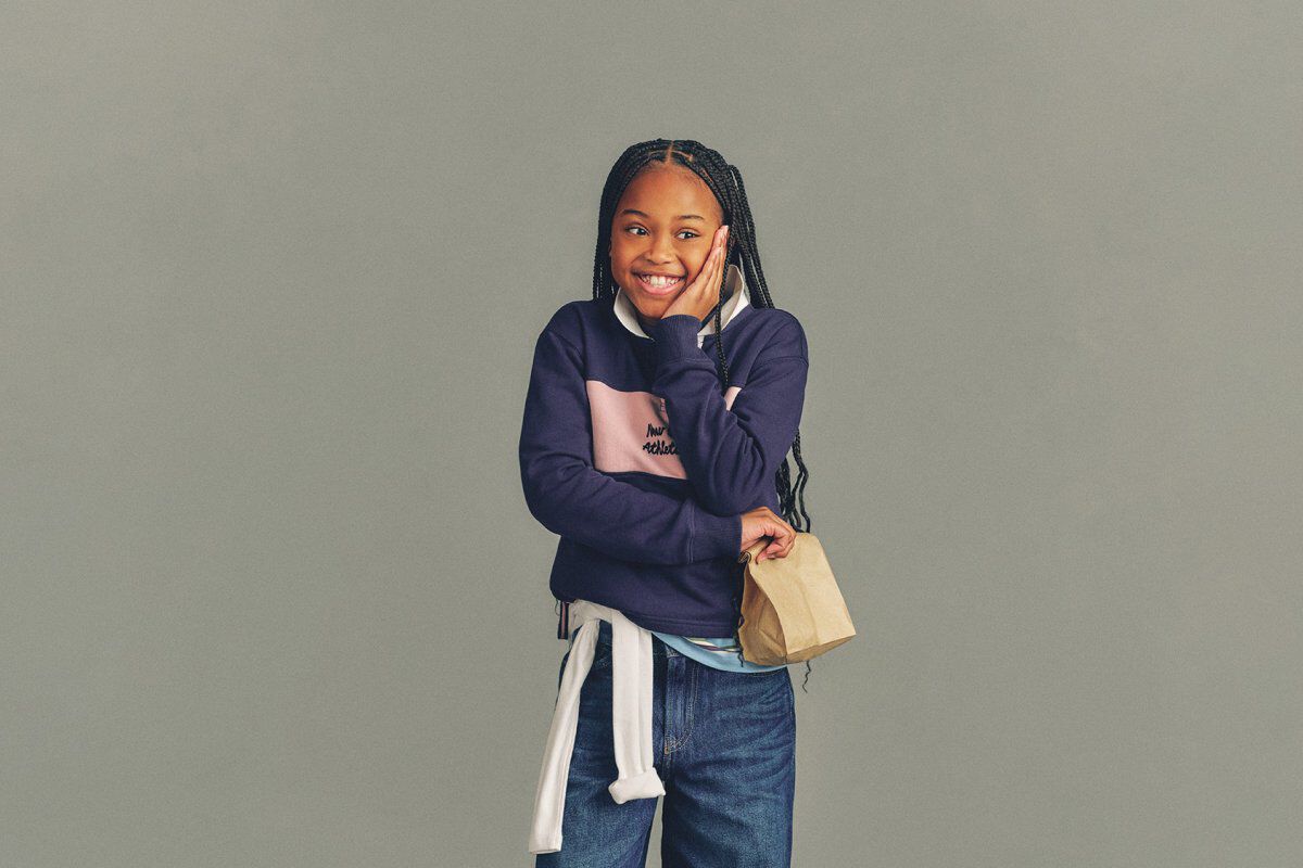 The child is wearing a light gray T-shirt layered under a soft pink zip-up hoodie with a white New Balance logo on the chest.