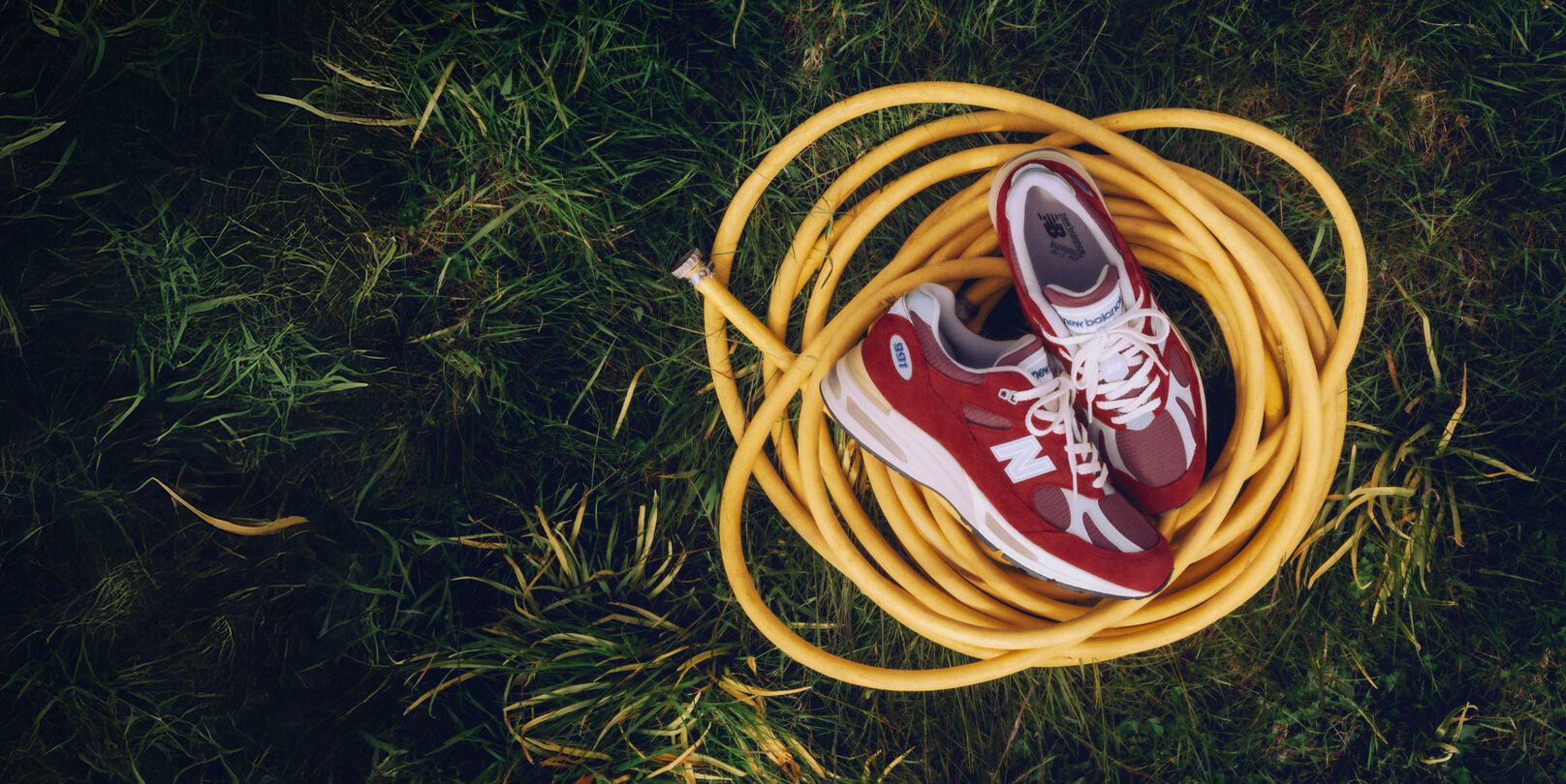 A pair of New Balance Made in UK sneakers sitting on a yellow coiled hose on top of a lush grass lawn. 