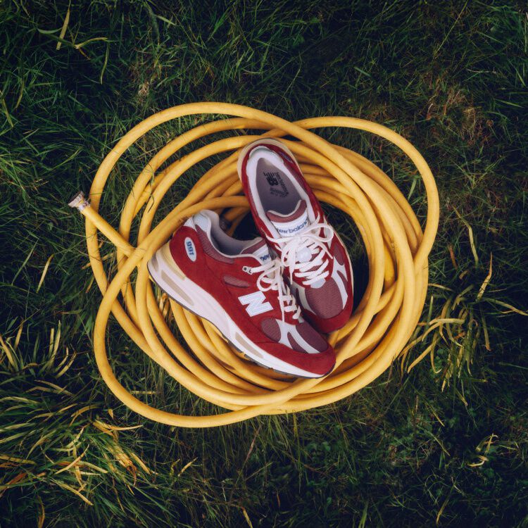 A pair of New Balance Made in UK sneakers sitting on a yellow coiled hose on top of a lush grass lawn. 